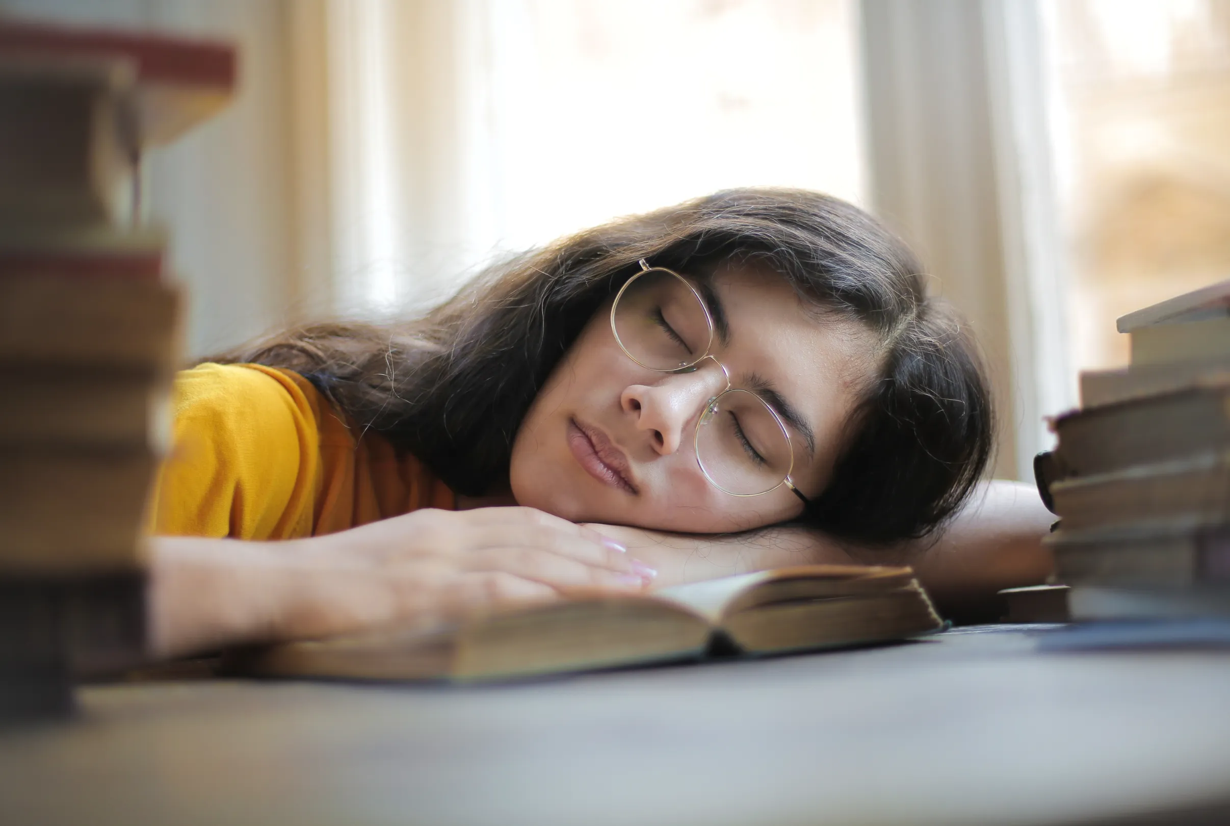 A young woman resting her head on an open book at a sunlit desk, eyes closed.