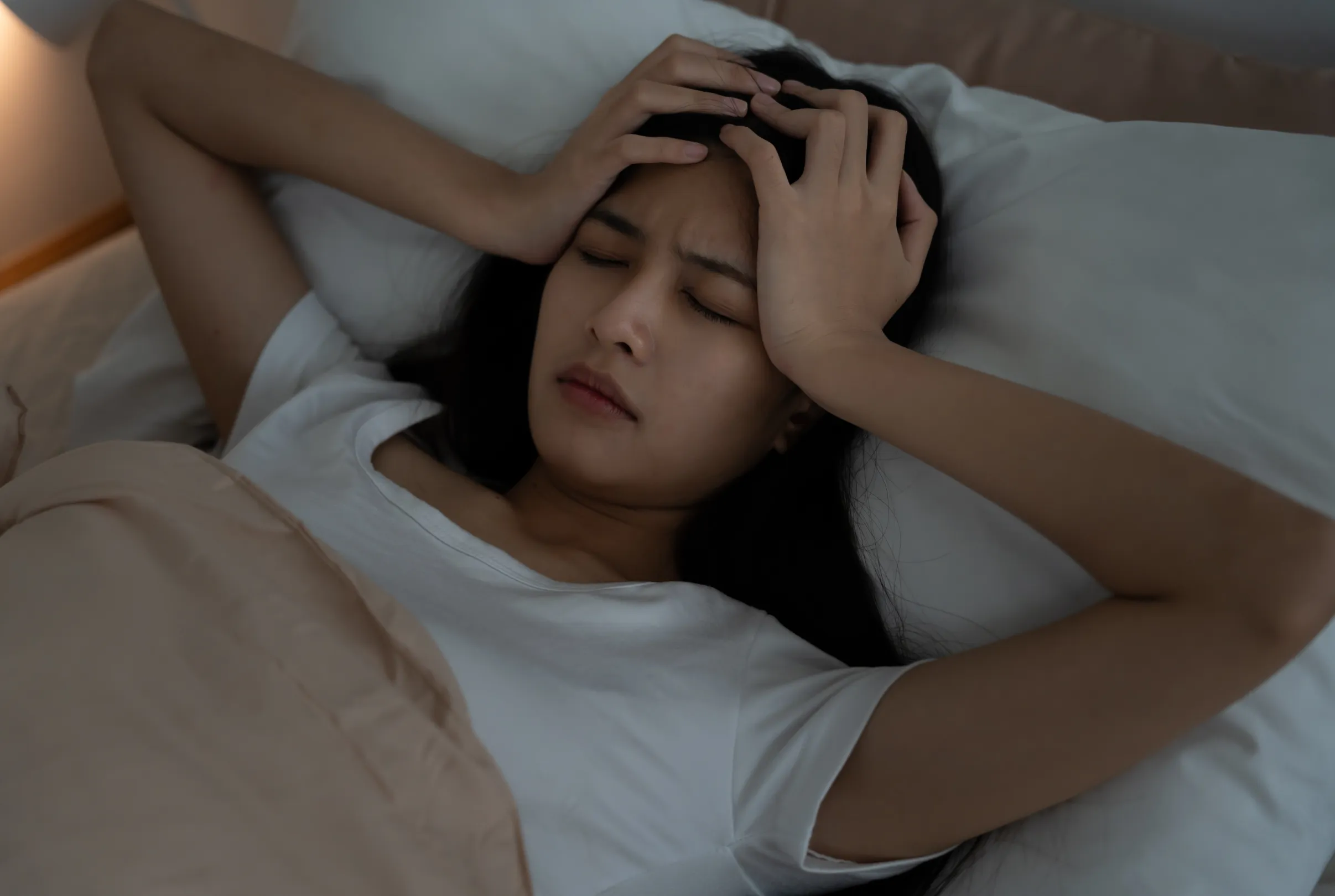 A woman lying in bed in a dimly lit room with both hands pressed against her forehead.