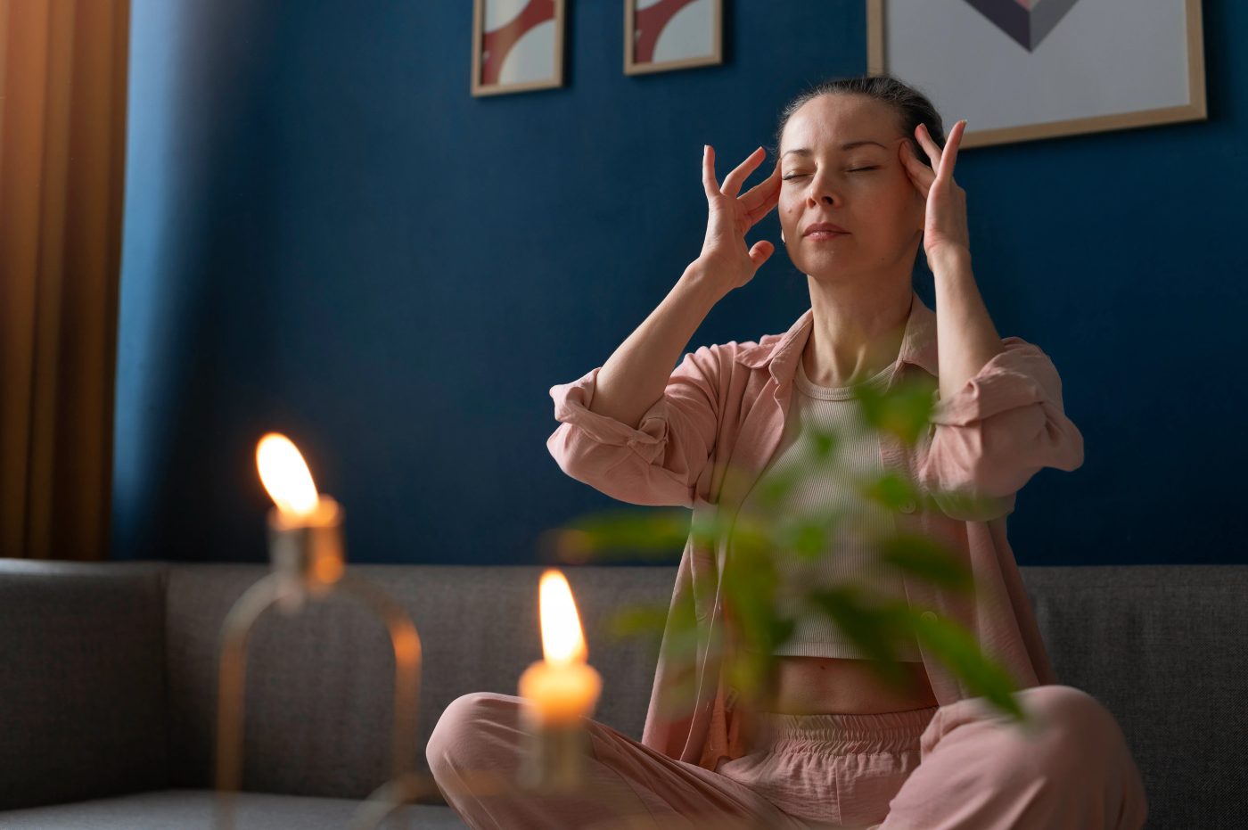 A woman sitting cross-legged on a sofa with eyes closed and fingertips at her temples, candles lit beside her.