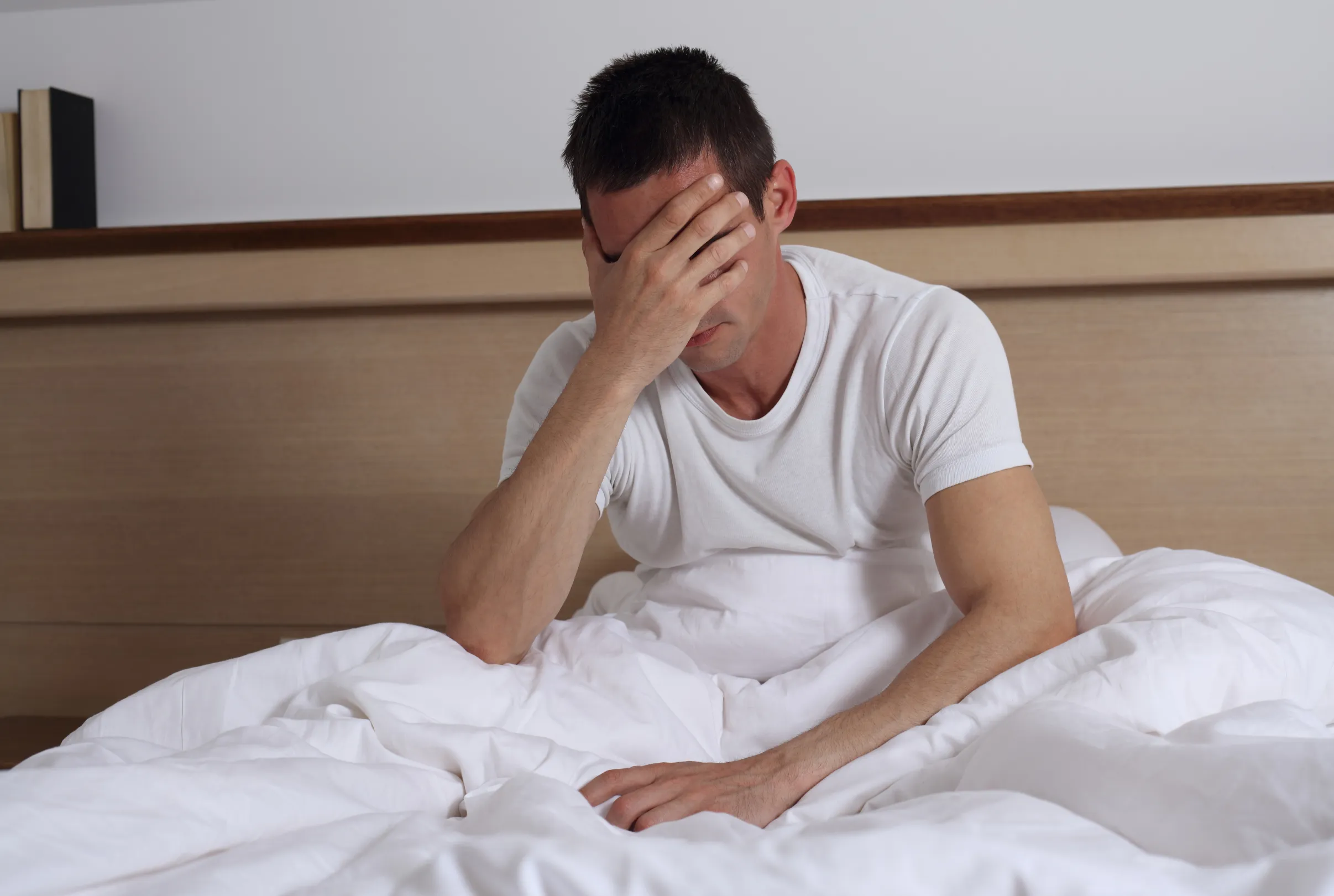 A man sitting up on the edge of his bed with one hand covering his face.